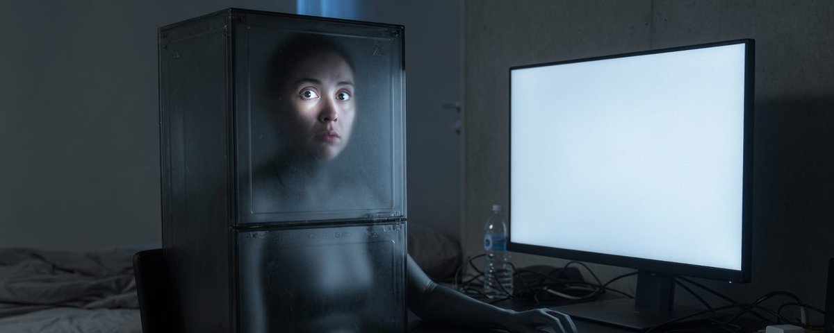 A woman sealed inside a glass box beside a glowing monitor
