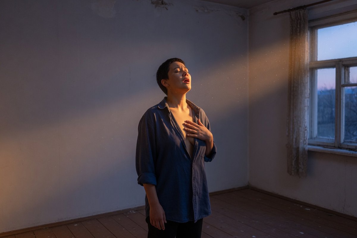 A woman standing in a bare room, hand on her chest, breathing in the light